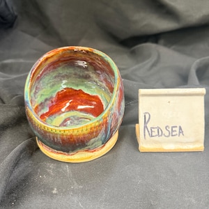 Puede incluir: Un cuenco de cerámica hecho a mano con un vibrante esmalte multicolor. El cuenco presenta tonos de rojo, verde y amarillo, con la palabra "REDSEA" escrita en una pequeña etiqueta. El cuenco tiene forma redondeada y superficie texturizada.