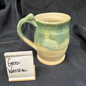 May include: A ceramic mug with a curved handle and a two-tone glaze featuring a glossy green drip pattern over a light beige base. Next to the mug is a small rectangular ceramic sign with the handwritten text Green Waterfall in black ink.