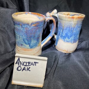 May include: Two handmade ceramic mugs with blue, brown, and cream glaze. The mugs have unique handles and a textured design. A small white tile with the words "Ancient Oak" written in blue ink is in front of the mugs.