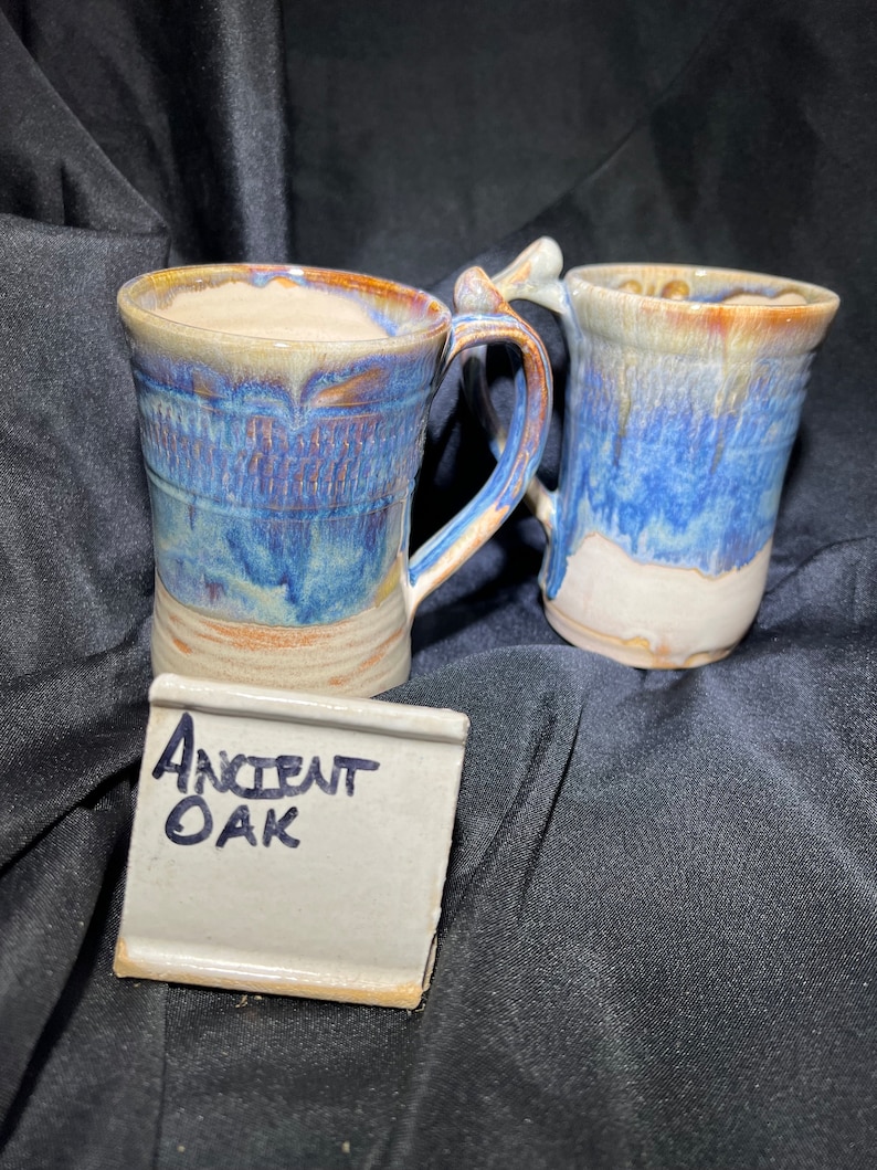 May include: Two handmade ceramic mugs with blue, brown, and cream glaze. The mugs have unique handles and a textured design. A small white tile with the words "Ancient Oak" written in blue ink is in front of the mugs.