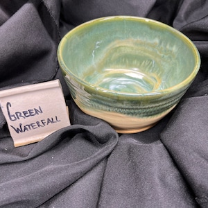 May include: A handmade ceramic bowl with a green and tan glaze. The bowl has a rounded shape and is on a black surface. A small tag next to the bowl reads "Green Waterfall".