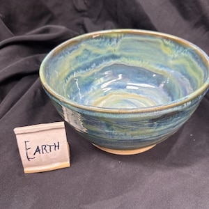 May include: A round ceramic bowl with a glossy glaze featuring swirling patterns of blue, green, and tan. Next to the bowl is a small, light-colored rectangular sign with a tan base that has the word EARTH written in black ink on its front surface.