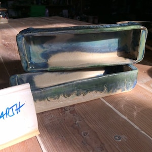 May include: Two rectangular ceramic trays stacked vertically, featuring a smooth beige base with a reactive glaze in shades of deep blue and forest green along the upper edges and interior. A small white ceramic tag with the blue text ARTH is visible.