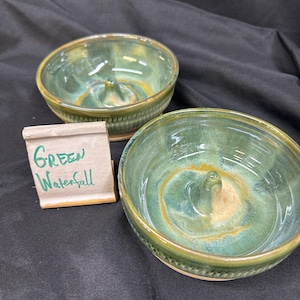May include: Two round ceramic ring dishes with a glossy green and amber glaze, each featuring a central cone. A small rectangular sign stands next to them, displaying the handwritten text Green Waterfall in green ink on a light beige background.