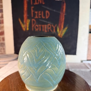 May include: A rounded ceramic vase with a light blue-green glaze and a carved chevron pattern. The vase has a smooth, glossy finish and a narrow opening at the top. The surface features a repeating V-shaped texture that wraps around the body.