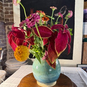 May include: A round, light blue and teal ceramic vase holds a bouquet of vibrant flowers and foliage. The arrangement features deep magenta coleus leaves with yellow edges, bright pink zinnias, and a single prominent golden-yellow zinnia.
