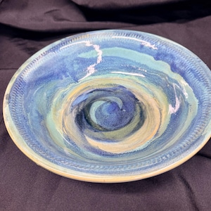 Puede incluir: Un cuenco de cerámica redondo con un patrón de esmalte en espiral en tonos azul marino profundo, azul cielo claro y crema suave. La superficie interior tiene un acabado brillante con un borde texturizado, mostrando colores que se arremolinan.