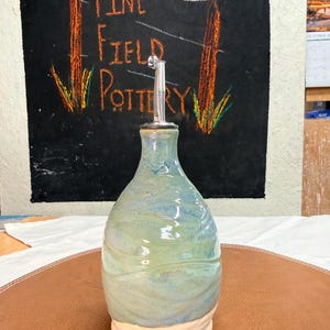 May include: A ceramic oil cruet with a glossy, mottled glaze in shades of blue and green, featuring a tapered body and a smooth, unglazed cream-colored base. The vessel is topped with a silver-colored metal pour spout insert.