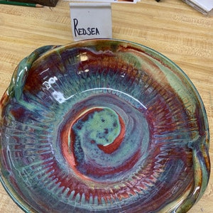 Könnte beinhalten: Eine große, handgefertigte Keramikschüssel mit einem einzigartigen Wirbeldesign in Rot-, Grün- und Brauntönen. Die Schüssel hat zwei Griffe und ist mit "REDSEA" beschriftet.