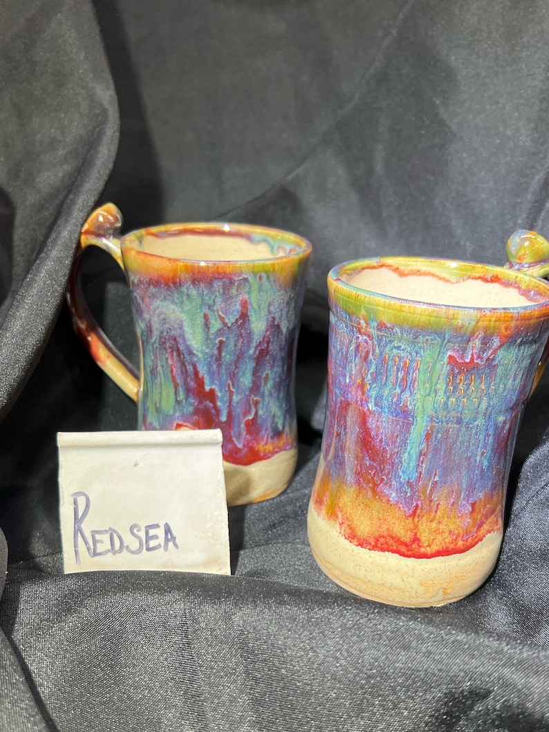 May include: Two handmade ceramic mugs with a unique glaze. The mugs feature a blend of red, blue, green, and yellow hues, with a beige base and a curved handle. The text "Redsea" is written on a small white card.