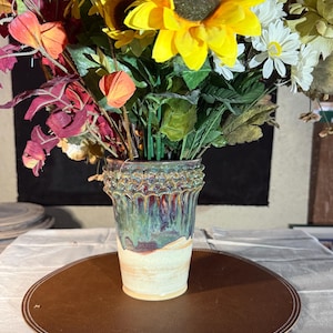 May include: A ceramic vase with a flared, scalloped rim and a tapered body. The upper portion features a glossy, dripped glaze in shades of deep green, blue, and purple, while the lower section has a smooth, matte cream-colored finish.
