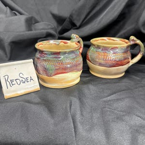 May include: Two ceramic mugs with a rounded shape and a handle, featuring a glaze of red, green, and tan colors with a textured band around the middle. A small white sign with the handwritten text "REDSEA" is placed next to the mugs.
