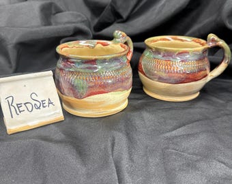 May include: Two ceramic mugs with a rounded shape and a handle, featuring a glaze of red, green, and tan colors with a textured band around the middle. A small white sign with the handwritten text "REDSEA" is placed next to the mugs.