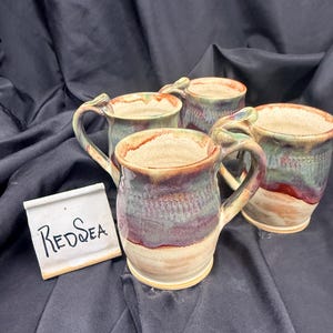 May include: Three ceramic mugs with a reactive glaze featuring blended tones of cream, sage green, and deep burgundy. Each mug has a curved handle and a textured body. A small white rectangular sign with the handwritten text RedSea is included.