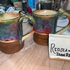 May include: Two handmade ceramic mugs with a textured base and a unique handle design. The mugs feature a blend of colors, including green, red, and blue, with a brown base. The text on the small white tile reads "Redsea w/ Iron Rim".
