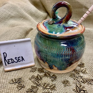Puede incluir: Un tarro de miel de cerámica hecho a mano con tapa y un cucharón de miel de madera. El tarro está esmaltado en tonos verdes, azules y marrones. La tapa tiene un asa de bucle y un pequeño dije de abeja. La palabra "Red Sea" está escrita en un pequeño letrero blanco.