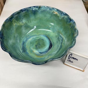 May include: A ceramic bowl with a wavy, scalloped rim featuring a glossy glaze in shades of blue and green. The interior displays a swirling, blended color pattern. A small white rectangular sign next to the bowl reads Green Sea.