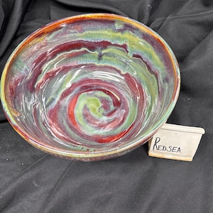 May include: A handcrafted ceramic bowl with a striking, swirling pattern in red, green, and yellow hues. The bowl features a glossy finish and an artistic aesthetic. The text "RED SEA" is on a small rectangular tag.