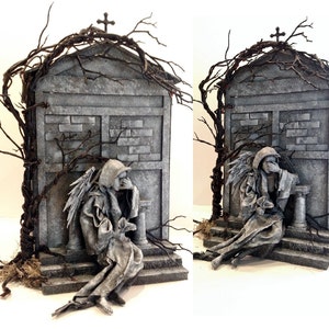 May include: A gray miniature mausoleum with a cross on top, adorned with dark branches. A seated angel sculpture with wings is positioned in front. The structure has a weathered, aged appearance, evoking a gothic or cemetery theme.