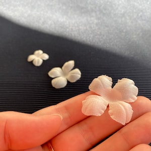 Könnte beinhalten: Drei weiße Stoffblumen, zwei kleine und eine große, mit einem fünfblattartigen Design. Die größere Blume wird in einer Hand gehalten.