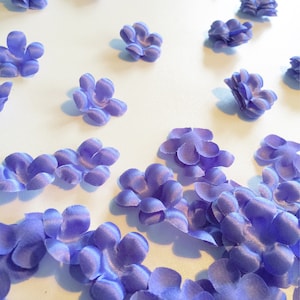 Puede incluir: Un surtido de pequeñas flores de tela de color lila claro dispersas sobre una superficie blanca. Las flores tienen múltiples pétalos y una apariencia ligeramente texturizada, lo que sugiere que están hechas de satén o un material similar. Ideales para manualidades o adornos.