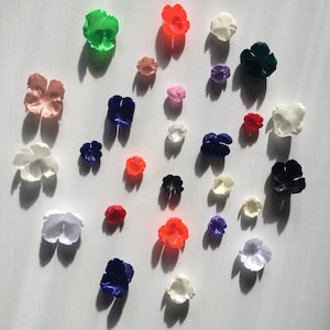 Puede incluir: Un surtido de pequeñas flores de tela coloridas en varios tonos, incluyendo rosa, blanco, rojo, azul y verde. Las flores están dispersas sobre una superficie blanca, proyectando sombras. Las flores son probablemente para manualidades o decoración.