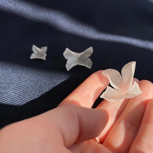 Könnte beinhalten: Drei weiße Stoffblumen, eine in einer Hand, zwei auf einer blauen Oberfläche. Die Blumen sind zart und haben eine weiche, luftige Textur.