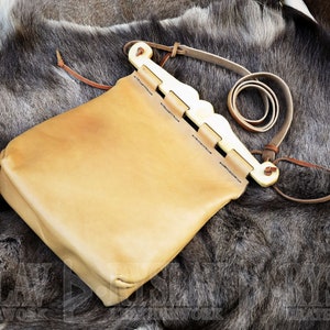 Könnte beinhalten: Eine hellbraune Ledertasche mit einem dekorativen, wellenförmigen Holzverschluss und einem dünnen Lederriemen. Die Tasche ist rechteckig mit abgerundeten Ecken und vor einem Hintergrund aus dunklem Fell.