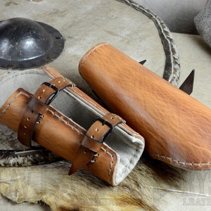 May include: Two brown leather arm guards with straps and buckles. The guards are lined with a light brown fabric. The guards are on a light brown fur surface.
