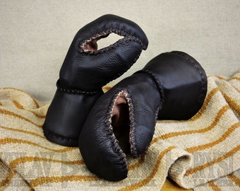 May include: A pair of black leather mittens. The mittens have a distinctive shape with a textured surface and contrasting stitching. The interior is a lighter brown. They are displayed on a striped beige and brown fabric.