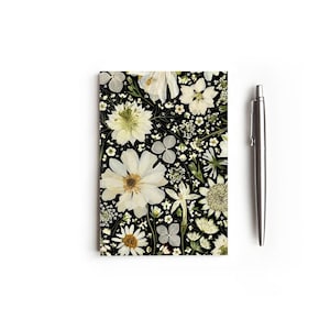 May include: A black notebook featuring a white floral pattern. The flowers are pressed and dried, creating a unique and delicate design.