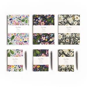 May include: Six notebooks with floral patterns, featuring pansies and other flowers, are displayed. Each notebook has a white band with the text "Lucy Miller Notebooks." Two silver pens are also visible.