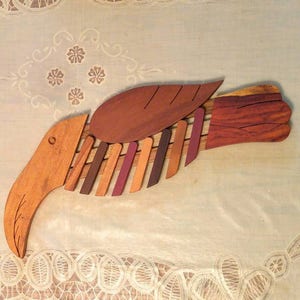 K&ouml;nnte beinhalten: Eine Holzskulptur eines Vogels mit einem langen Schnabel und einem Fl&uuml;gel aus mehreren Holzst&uuml;cken. Der Vogel ist in Braun-, Rot- und Lilat&ouml;nen bemalt.