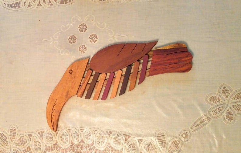 K&ouml;nnte beinhalten: Eine Holzskulptur eines Vogels mit einem langen Schnabel und einem Fl&uuml;gel aus mehreren Holzst&uuml;cken. Der Vogel ist in Braun-, Rot- und Lilat&ouml;nen bemalt.