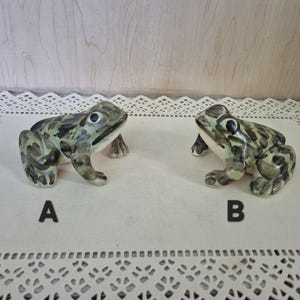 May include: Two ceramic frog figurines, labeled A and B, featuring a glossy finish with mottled green and brown spots. Each frog is depicted in a seated position with dark eyes, a light-colored underbelly, and detailed limbs resting on the surface.