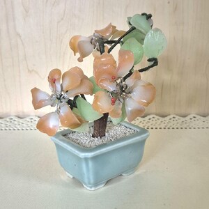 May include: A decorative bonsai tree with orange and white flower-shaped ornaments and green leaves. The tree is in a light blue square ceramic pot filled with small white stones. The background is a light wood panel.