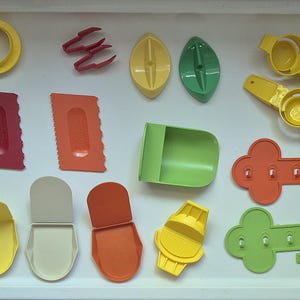 May include: A collection of colorful plastic kitchen tools, including egg separators, spatulas, and key-shaped wall hooks. The tools are in shades of yellow, red, orange, and green, arranged on a white tray.