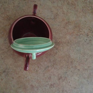 May include: A red ceramic teapot with a green lid. The lid is off and the teapot is sitting on a brown surface.
