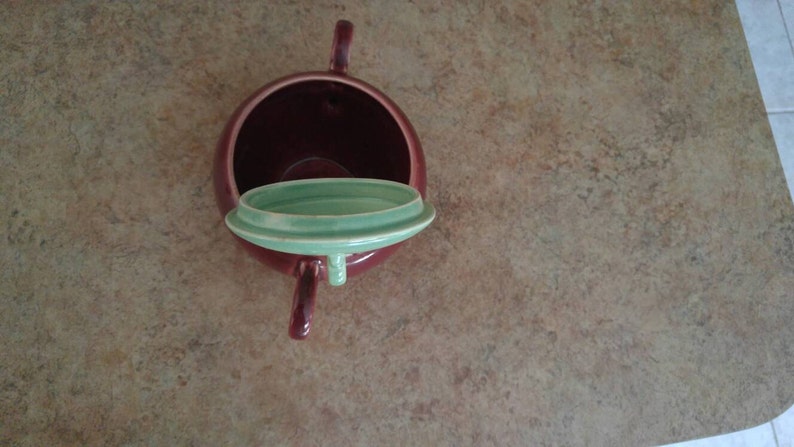 May include: A red ceramic teapot with a green lid. The lid is off and the teapot is sitting on a brown surface.