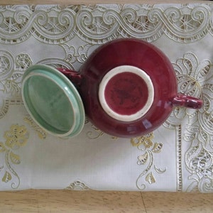 May include: A red ceramic teapot with a green lid. The teapot has a handle and a spout. The lid is slightly ajar.
