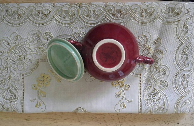 May include: A red ceramic teapot with a green lid. The teapot has a handle and a spout. The lid is slightly ajar.