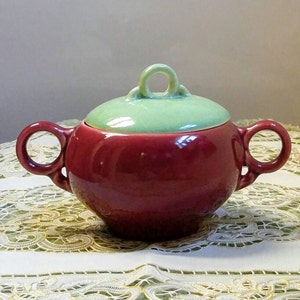 May include: A red ceramic sugar bowl with a green lid and two handles. The bowl has a rounded shape and a smooth finish.