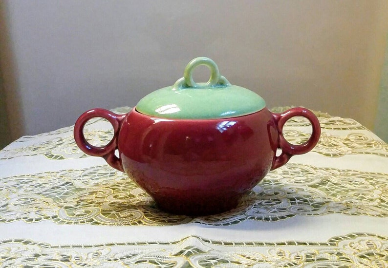 May include: A red ceramic sugar bowl with a green lid and two handles. The bowl has a rounded shape and a smooth finish.