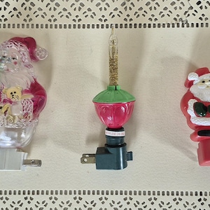 May include: Three Christmas-themed night lights. One features Santa Claus holding a tree and a teddy bear. Another is a red and green light with a gold glitter bulb. The third is a Santa Claus figure.