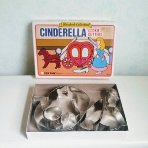 May include: A set of Cinderella cookie cutters in a white box with a colorful illustration of Cinderella, her carriage, and a glass slipper. The cutters are made of metal and include shapes like Cinderella, her carriage, a glass slipper, and a star.