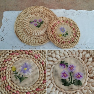 May include: Two round woven straw coasters with pink trim. Each coaster features a floral embroidery design with purple, pink, and green flowers.