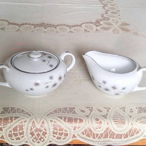 May include: A white porcelain creamer and sugar bowl set with a delicate floral design. The creamer has a silver rim and a handle. The sugar bowl has a lid with a silver knob.