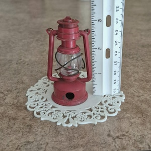 May include: A small, red metal lantern with a clear glass globe, standing on a white, decorative doily. The lantern is approximately 3 inches tall. The lantern has a handle and a small hole at the base.