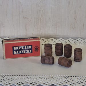 May include: A rectangular cardboard box with a red and blue label reading LIONEL ELECTRIC TRAINS and THE LIONEL CORPORATION, accompanied by six small, dark brown wooden barrel-shaped figures arranged in a cluster next to the box.