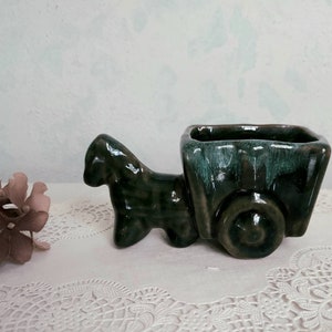 May include: A green ceramic planter shaped like a horse-drawn cart. The cart has a square planter box and a single wheel. The horse is standing on all four legs and has a detailed face.